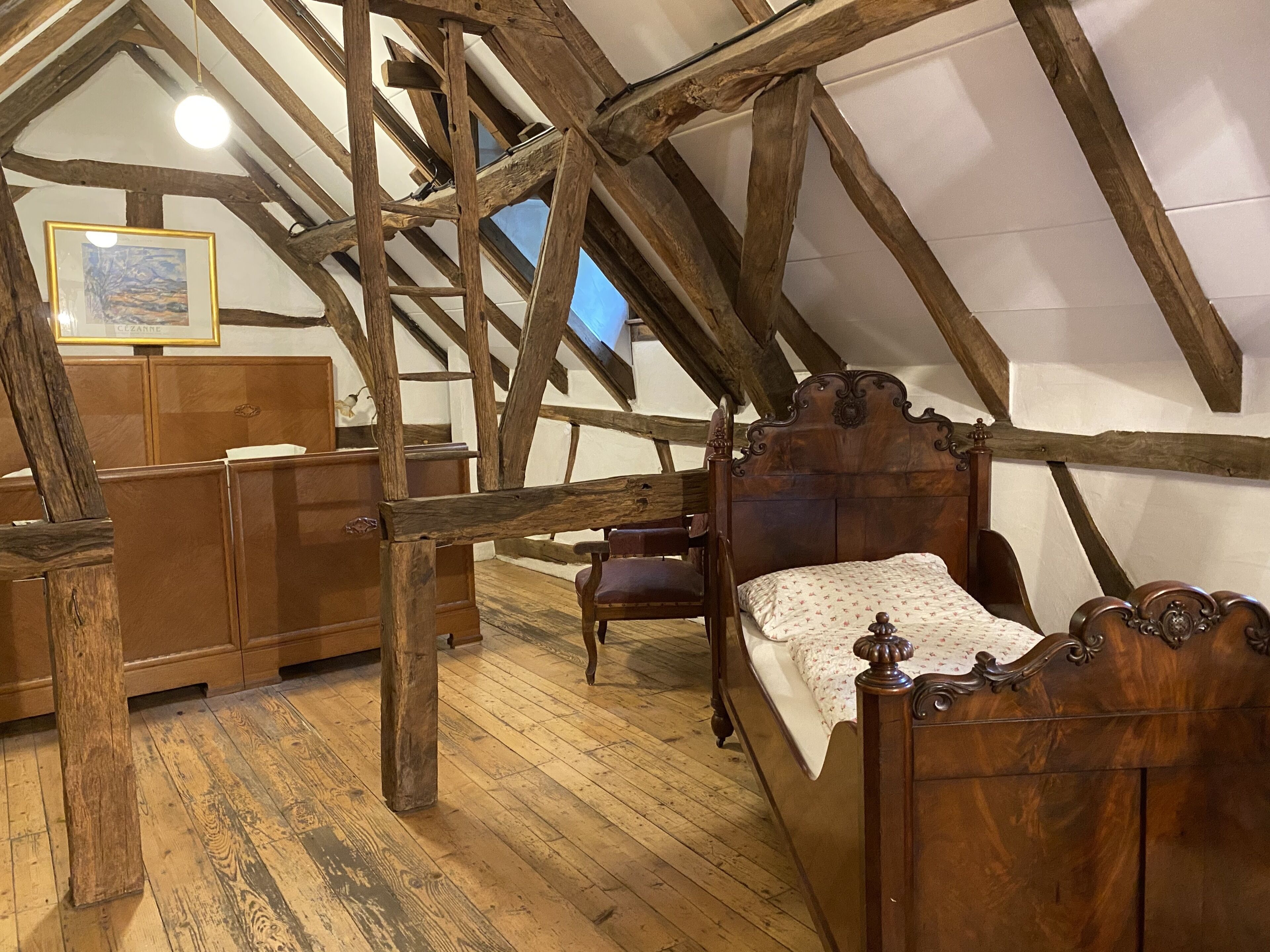 A rustic attic bedroom with exposed wooden beams, a wooden bed, a wooden chair, and a wooden dresser.