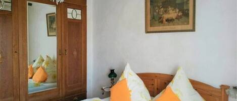 A bedroom with a wooden wardrobe, a bed with orange and white pillows, a green lamp, and a framed painting on the wall.
