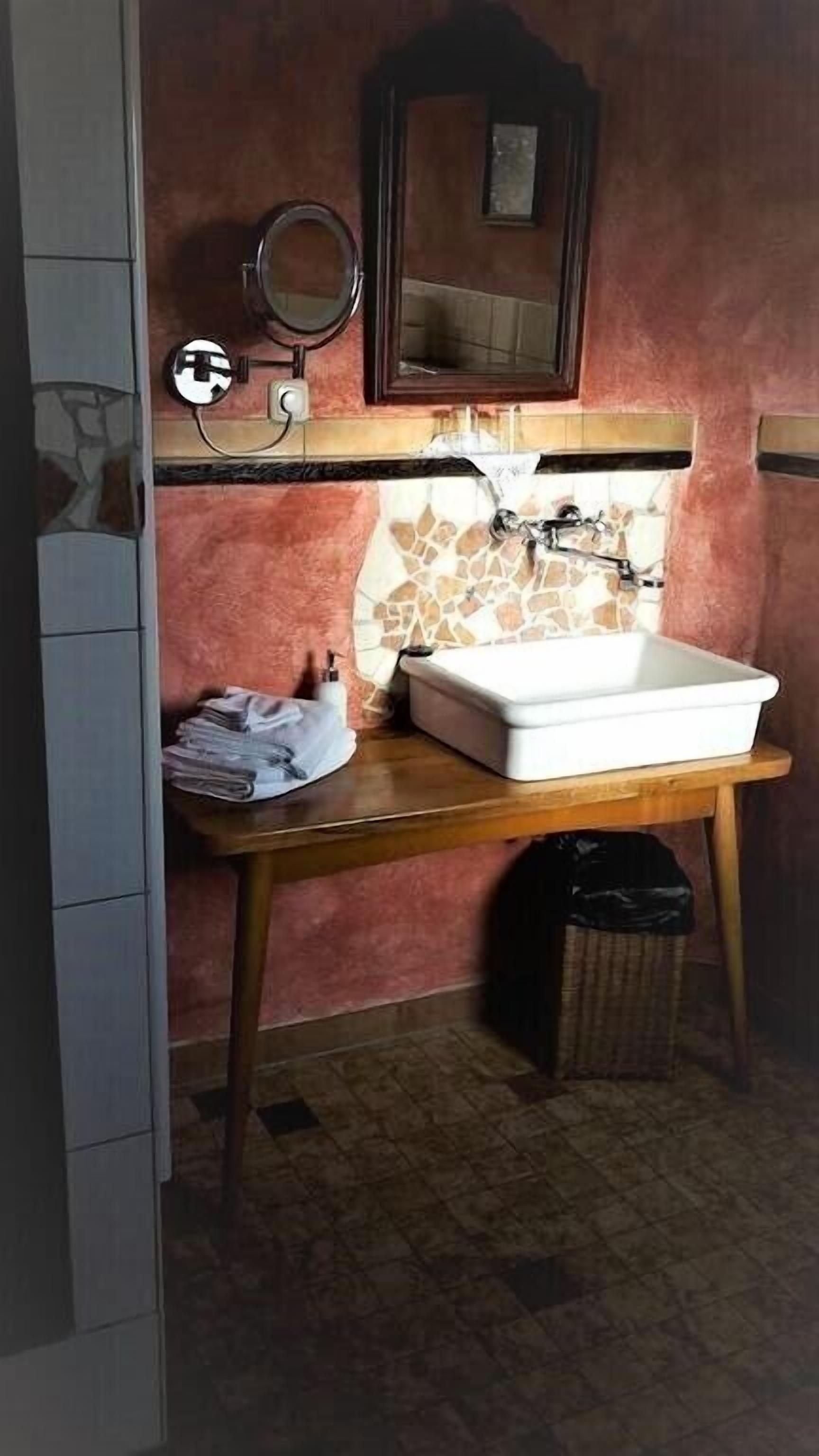 A bathroom with a white sink, a mirror, and a tiled backsplash. There is a trash bin and a folded towel on the counter.