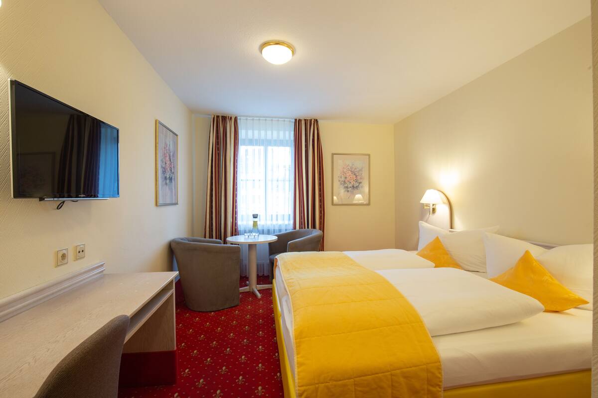 Habitación de hotel con cama, escritorio, silla, televisión y ventana con cortinas.