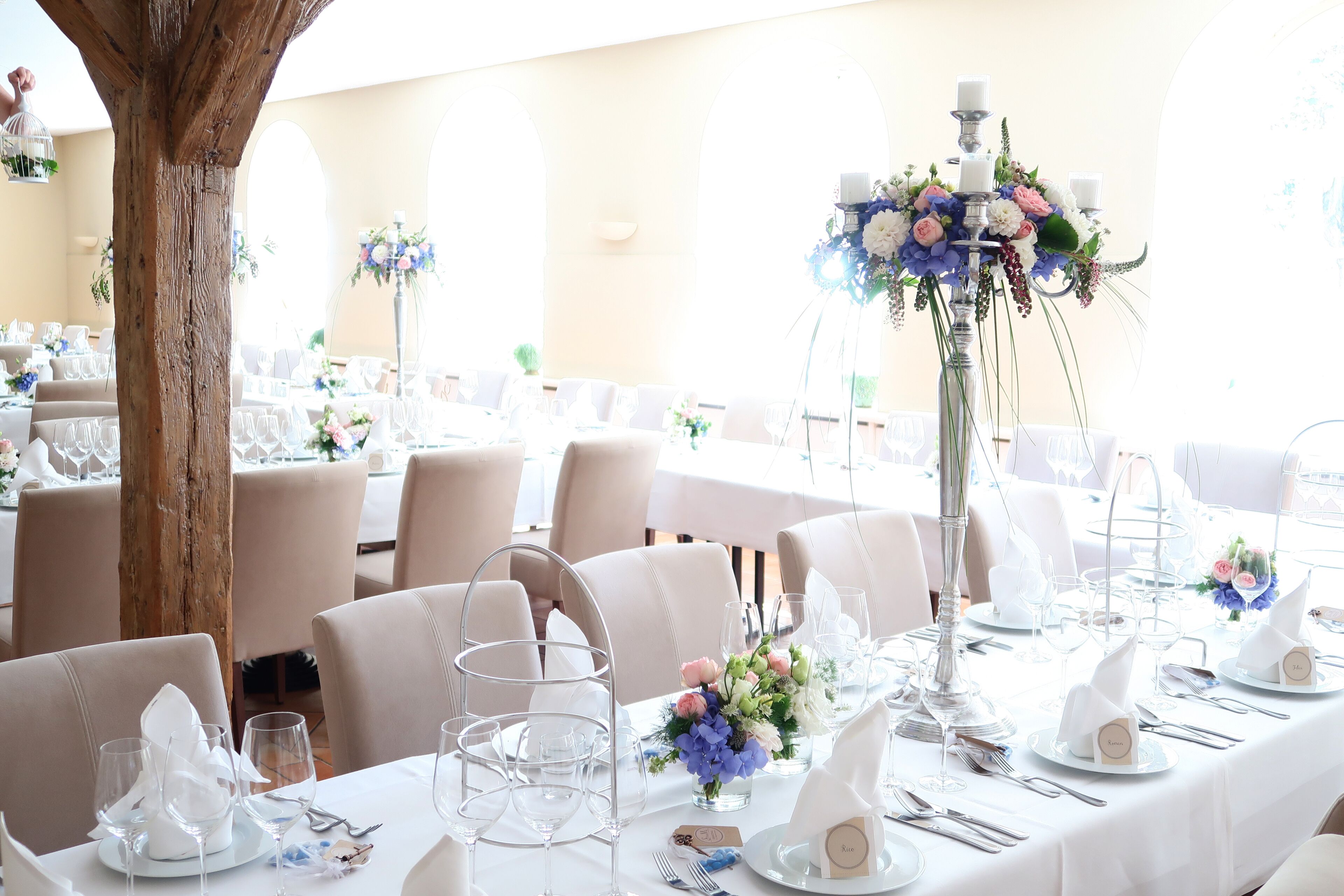 Uma sala de banquetes com mesas redondas preparadas para um evento formal, com toalhas brancas, copos de vidro e arranjos florais.