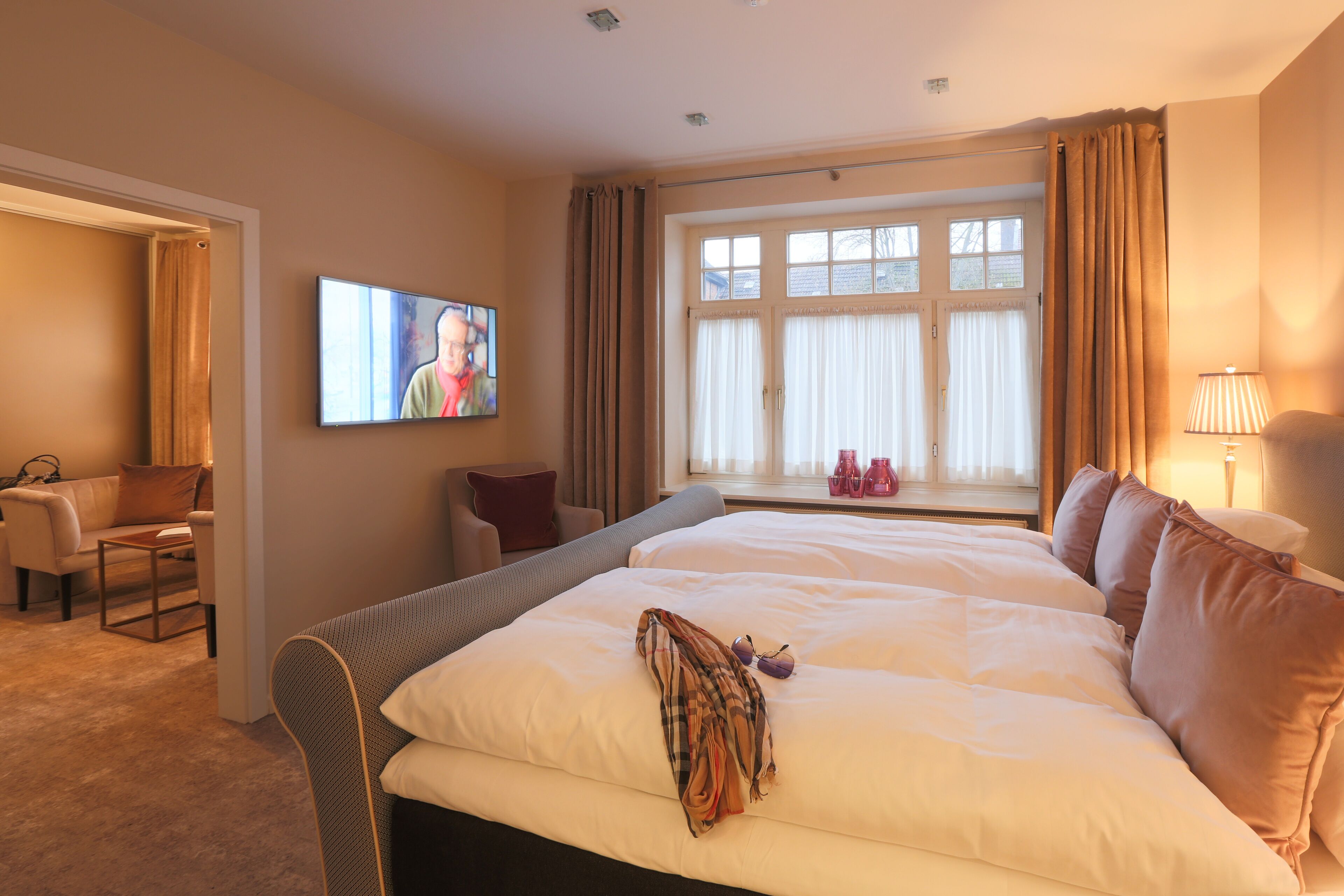 Habitación de hotel con una cama grande, un televisor en la pared, una ventana con cortinas, una silla y una lámpara.
