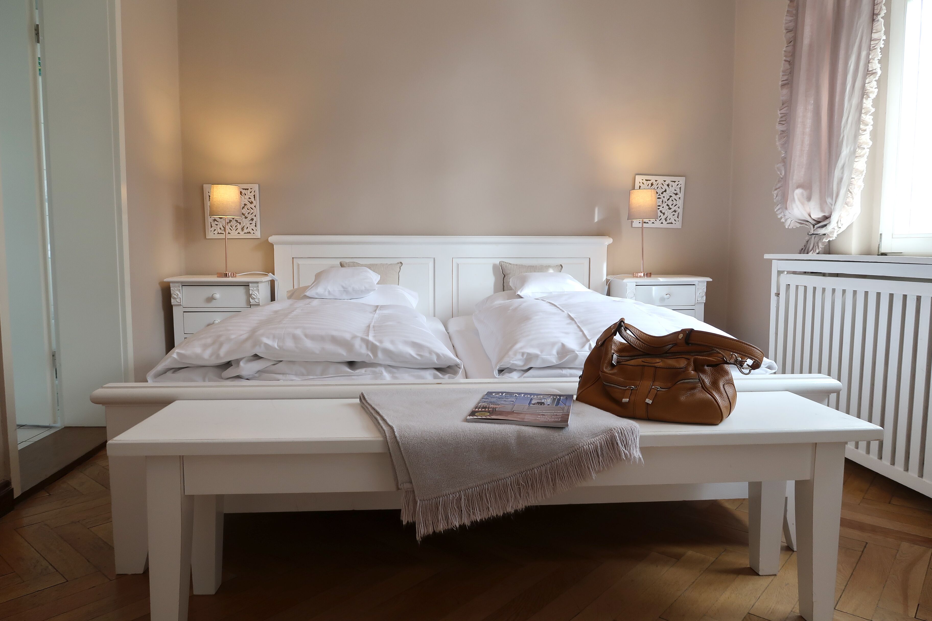 A neatly made double bed with white linens, two bedside tables with lamps, a bench with a magazine and a handbag, and a window with curtains.