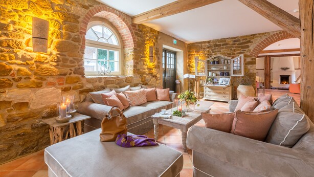 Una habitación acogedora con paredes de piedra, techo de madera y chimenea. Se observan un sofá, una mesa baja y una mesa de comedor.