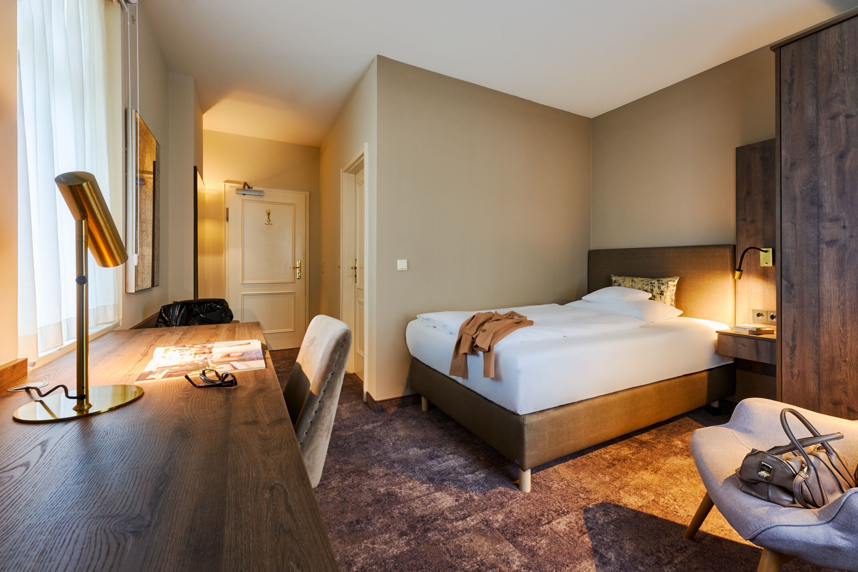 Una habitación de hotel con cama, mesita de noche, lámpara, silla y un escritorio con otra silla.