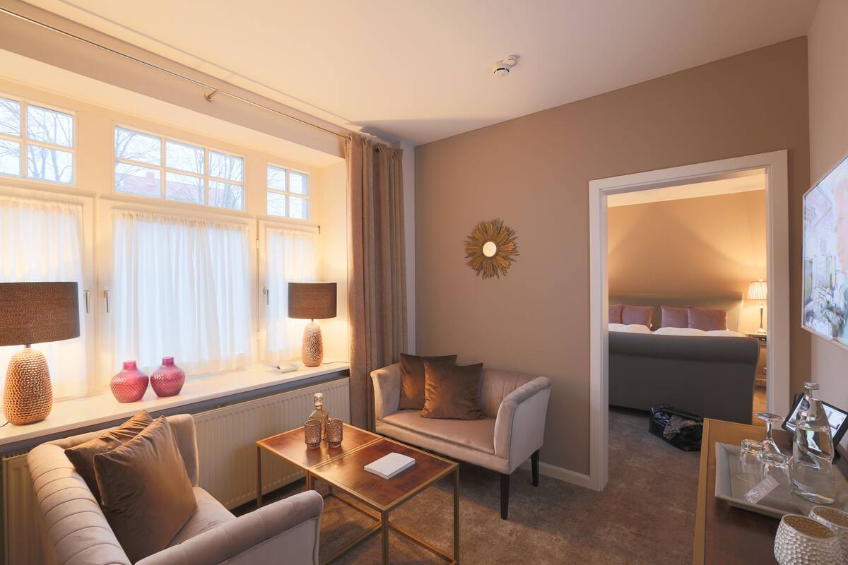 A hotel room with a sofa, armchair, and a small table. There is a window with curtains, a radiator, and a wall-mounted lamp.