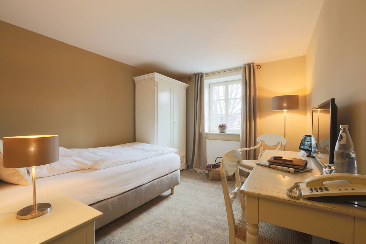A hotel room with a bed, a desk with a lamp, a chair, a telephone, a bottle, and a window with curtains.