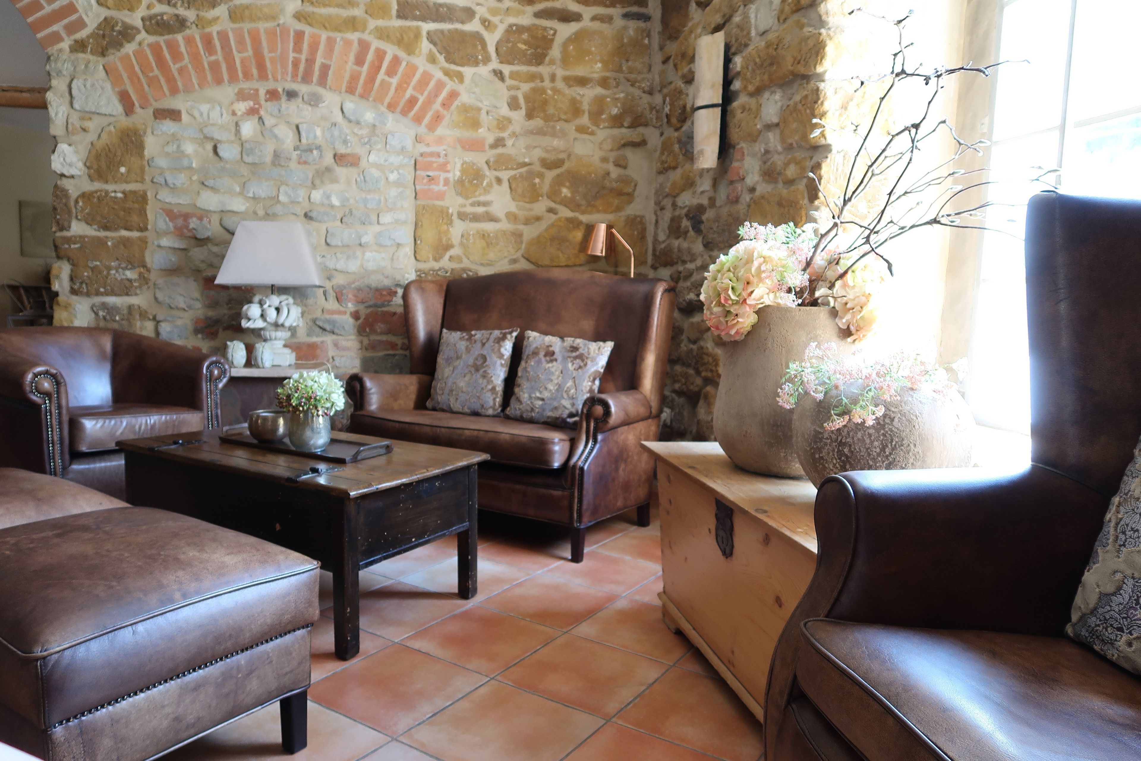 Una sala de estar acogedora con pared de piedra, mobiliario de cuero y un jarrón grande con flores.