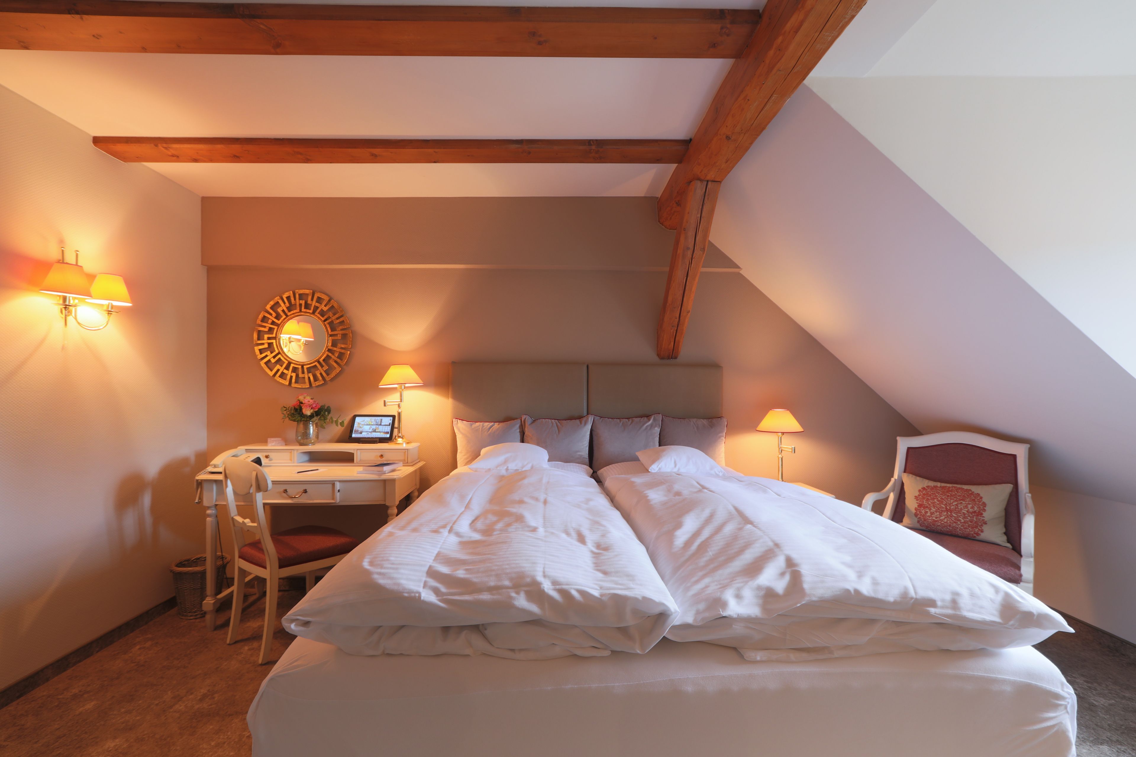 Un dormitorio con una cama grande, un escritorio y dos sillas. Hay apliques y un espejo redondo en la pared.