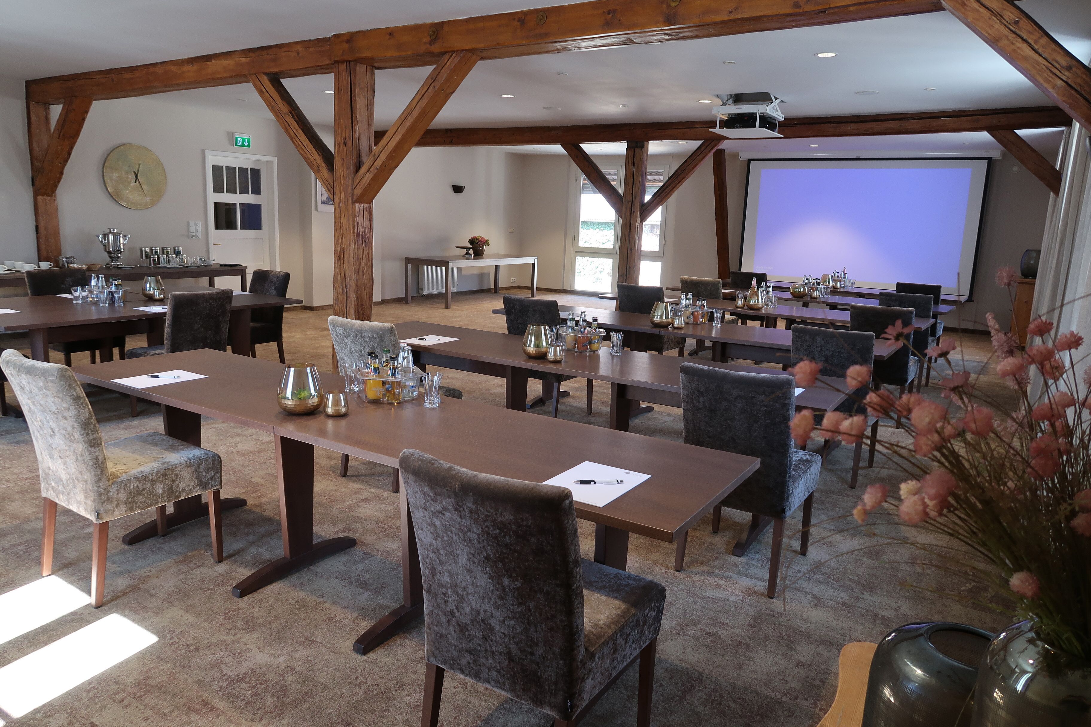 Une salle de conférence dotée de plusieurs tables et chaises, de poutres en bois au plafond, d’un écran de projection et d’une horloge au mur.