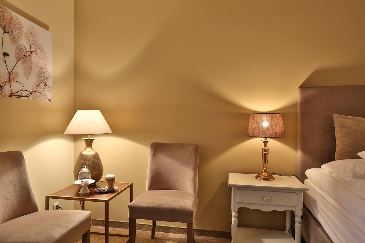 A hotel room with a bed, two chairs, a small table with a lamp, and a nightstand with a lamp.