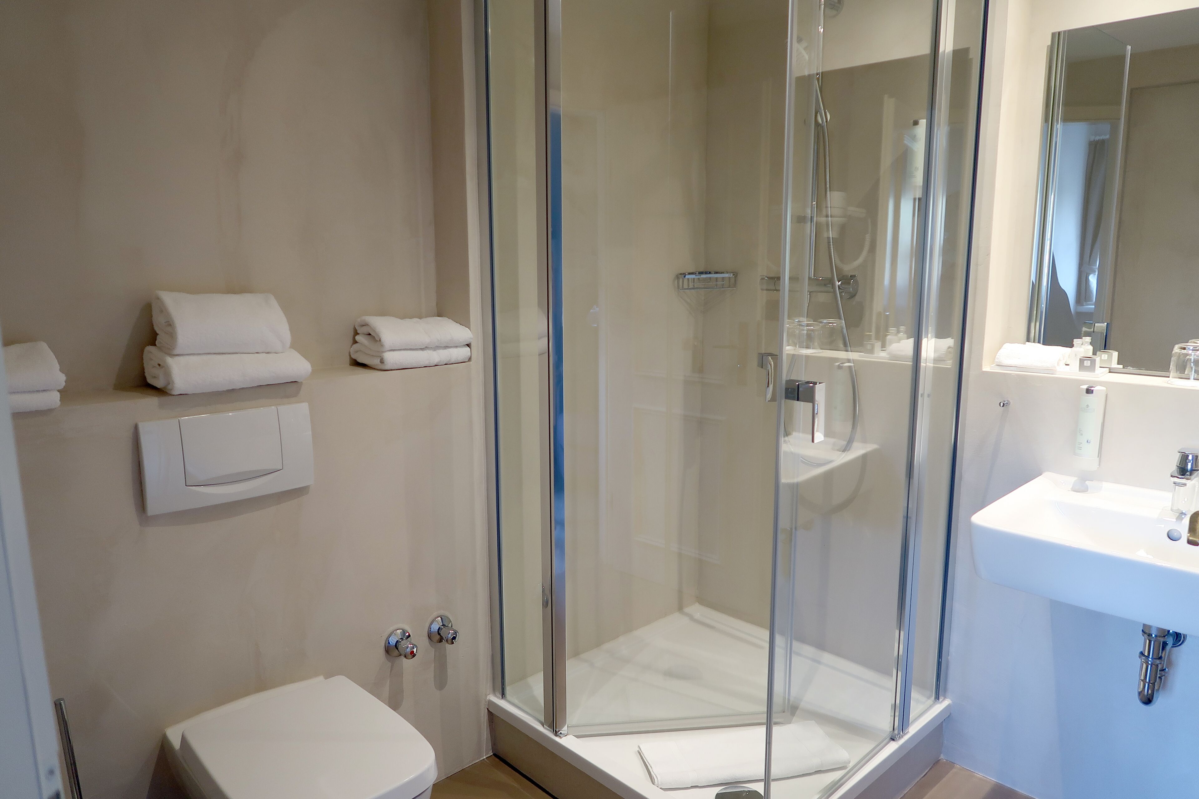 Un baño moderno con ducha de vidrio, un lavabo blanco y toallas ordenadas sobre una repisa.