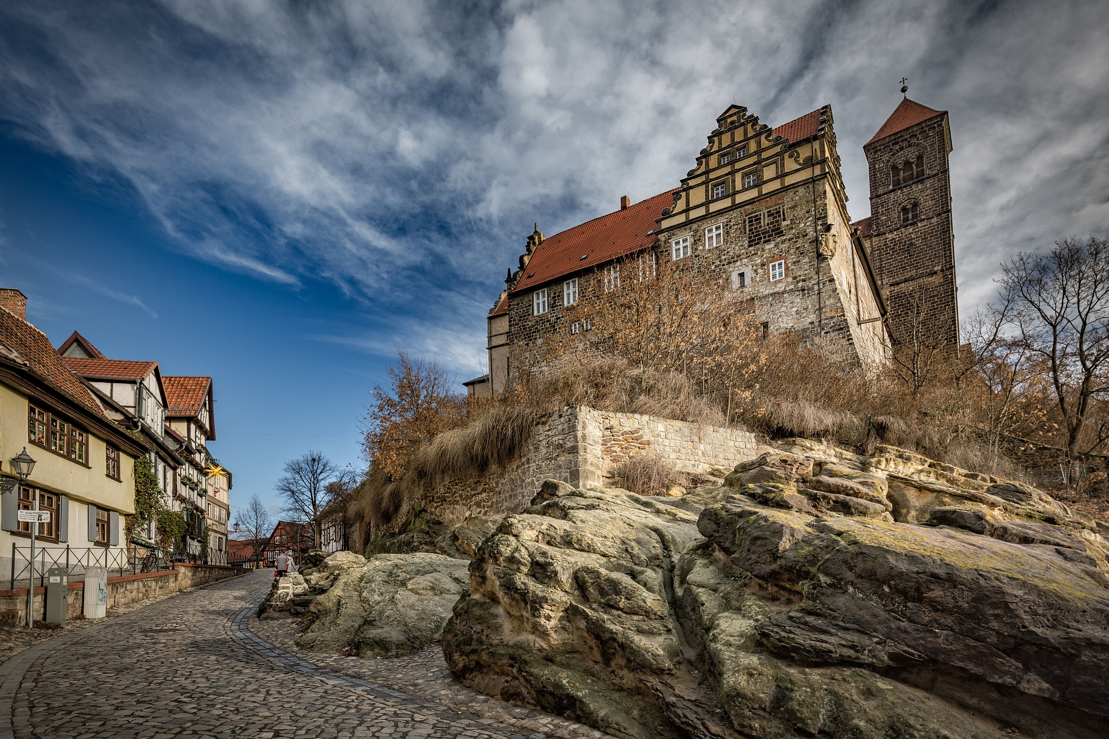 A historic stone building with a red-tiled roof, situated on a cobblestone street with large rocks in the foreground.