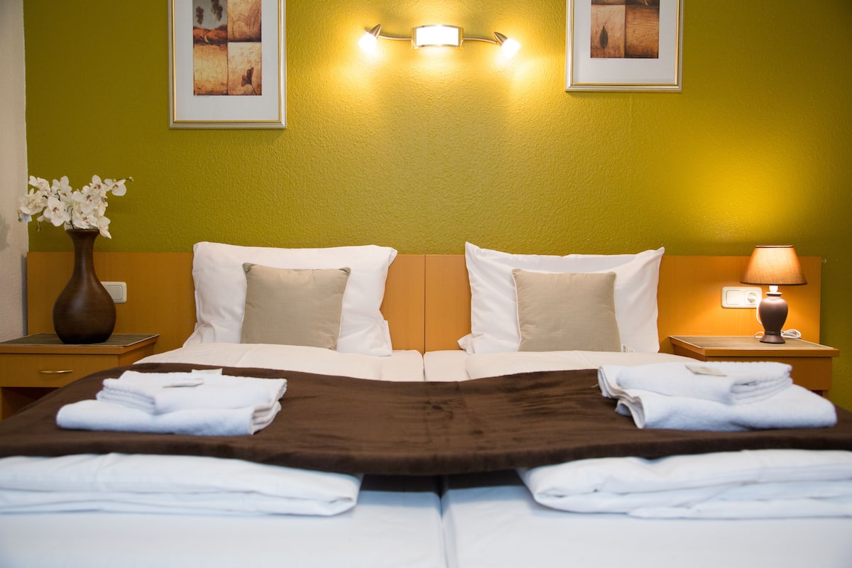 Een hotelkamer met twee bedden, een bruin hoofdbord, wit beddengoed, een vaas met bloemen en een nachtkastje met een lamp.