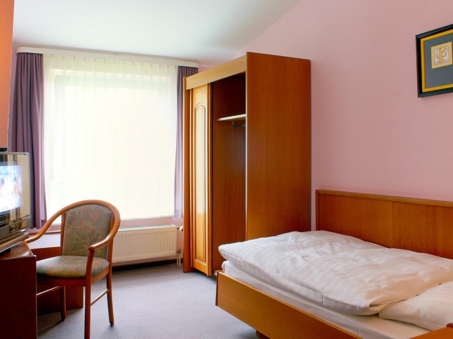 A hotel room with a bed, a chair, a desk, and a radiator.
