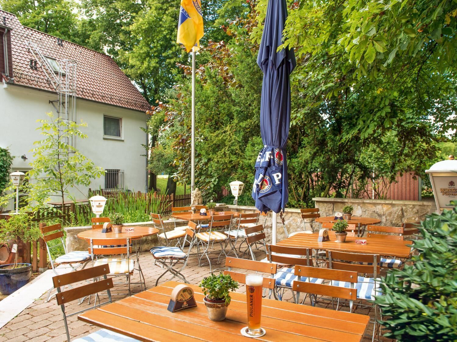 An outdoor patio with wooden tables and chairs, surrounded by greenery and a building with a tiled roof.