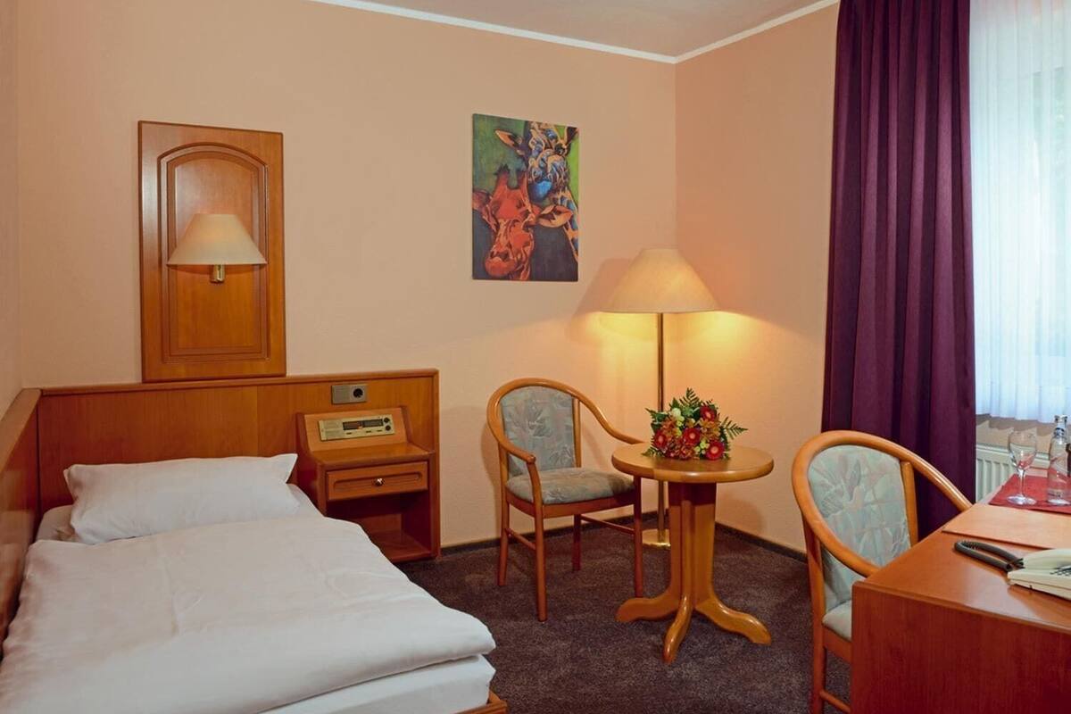 Habitación de hotel con una cama, dos sillones, una mesita de noche con lámpara, un cuadro en la pared y una ventana con cortinas.