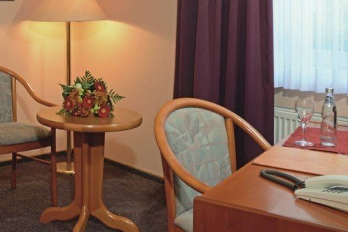 Habitación de hotel con cama, dos sillas, una mesita con lámpara, escritorio con teléfono y ventana con cortinas.