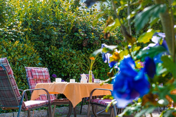 An outdoor dining area with a table set for two, surrounded by lush greenery and a hedge.