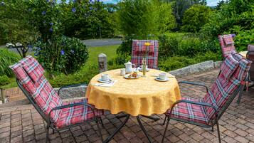 A round table with a yellow tablecloth, surrounded by plaid chairs, set outdoors with a garden view.