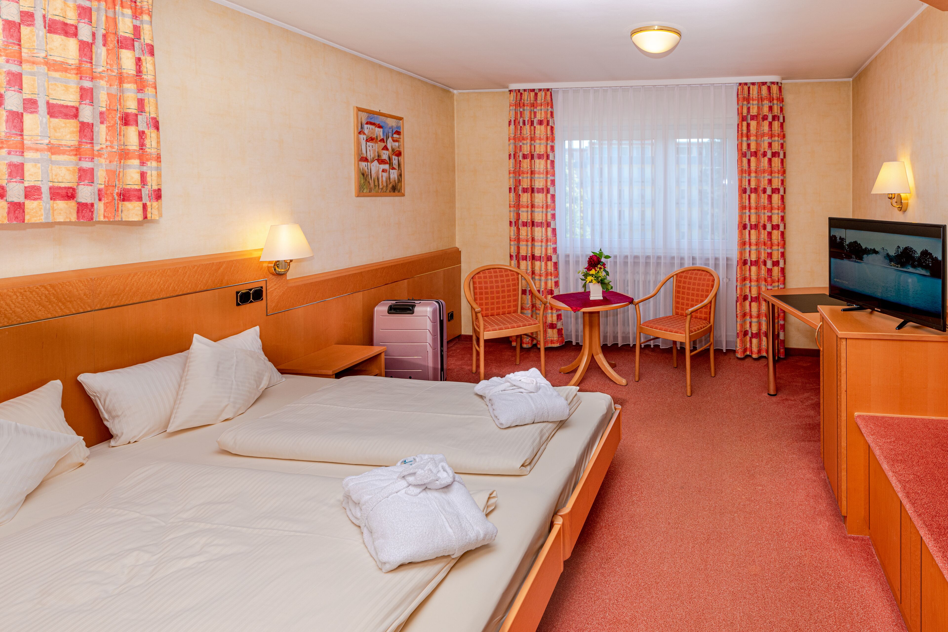 A hotel room with two beds, a desk, a television, a chair, and a small table with a flower vase.