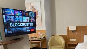 A hotel room with a television displaying the 'Blockbuster' channel, a desk, a chair, a bed, and a small stool. - Gästehaus Kral (Erlangen)