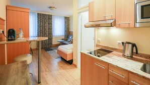 A compact kitchen with a microwave, stove, and sink. There is a small dining area with a table and chairs. The room has wooden flooring and a window with curtains. - Gästehaus Kral (Erlangen)