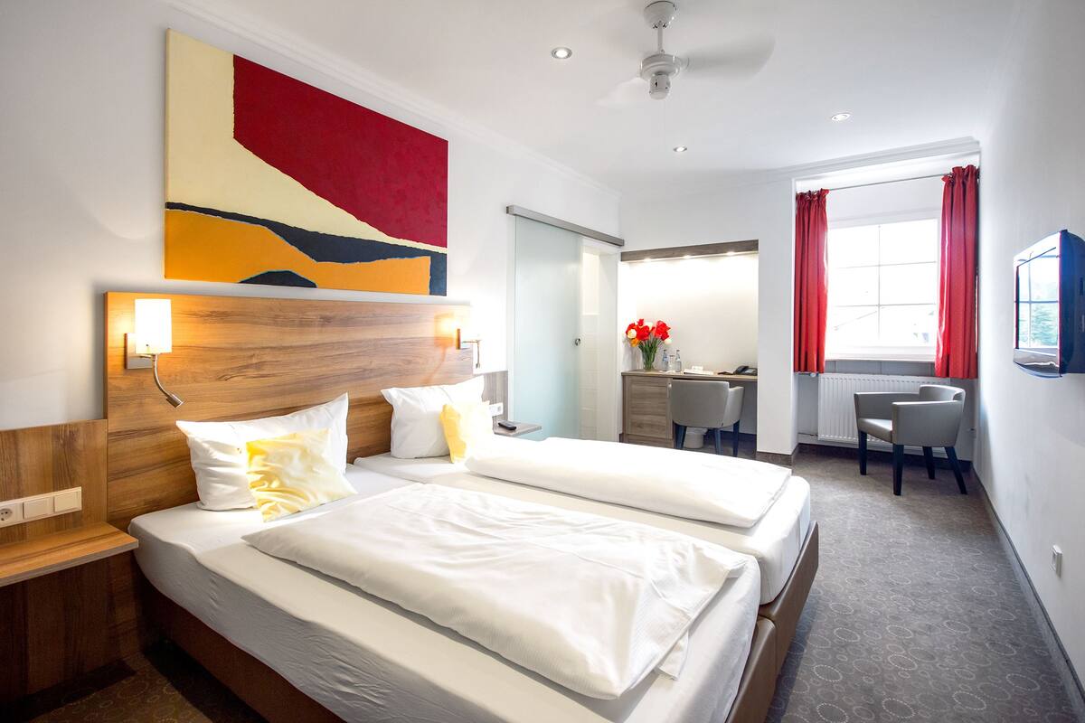 Habitación de hotel con dos camas, un escritorio, una silla, un cuadro abstracto grande y una ventana con cortinas rojas.