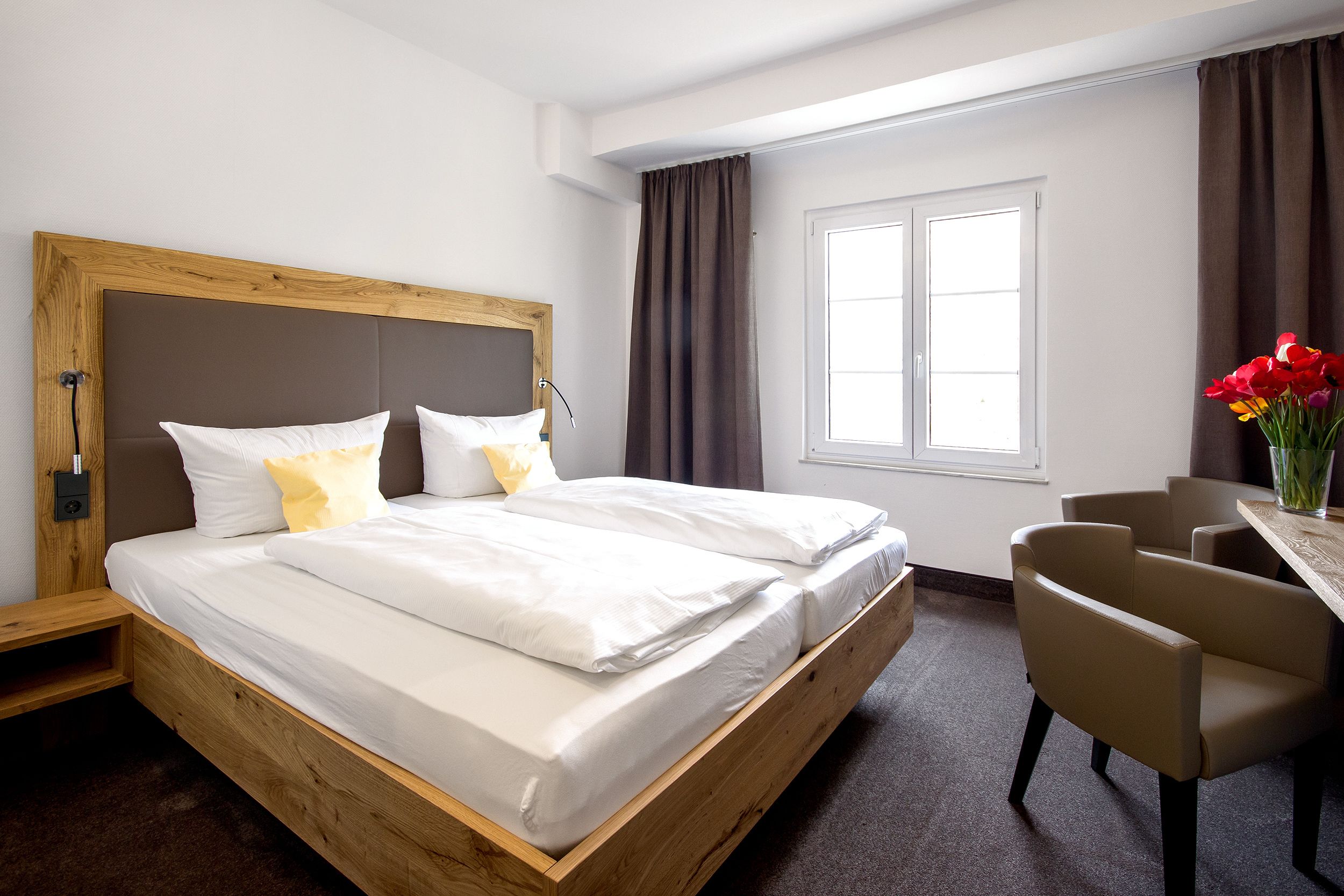 A modern hotel room with a wooden bed frame, white bedding, a desk with a chair, and a window with curtains.