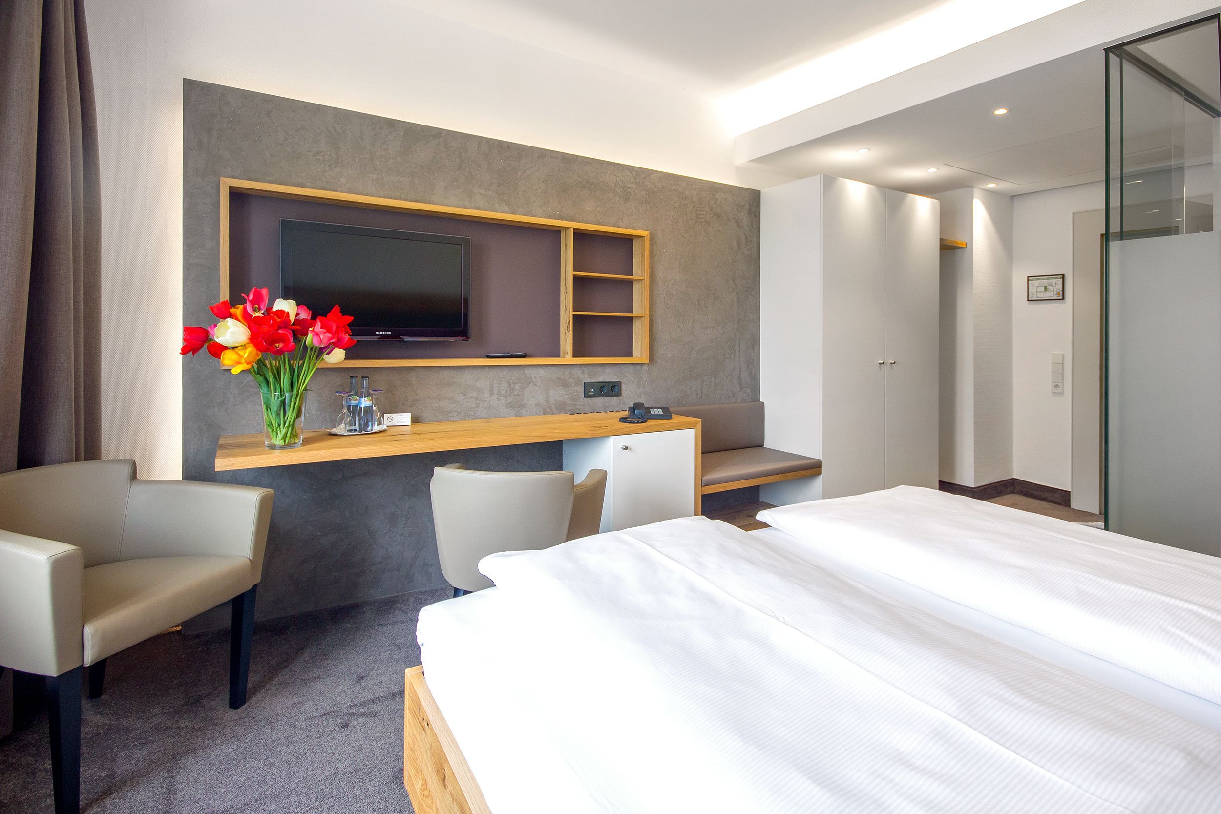 Une chambre d’hôtel comprenant un lit, deux chaises, un bureau avec un téléviseur à écran plat, un vase avec des fleurs et une armoire.