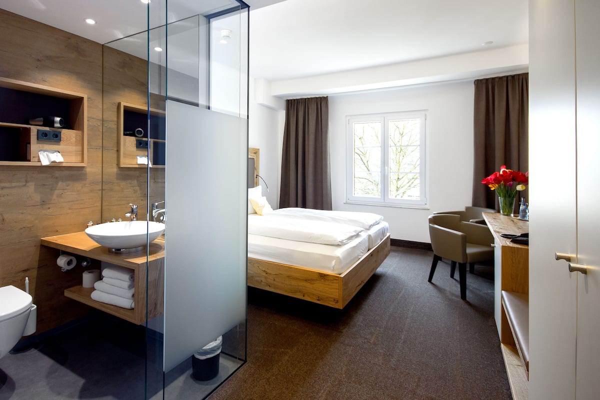 Una habitación de hotel moderna con una cama de madera, un baño con paredes de vidrio y un escritorio con silla, además de un ventanal con cortinas.