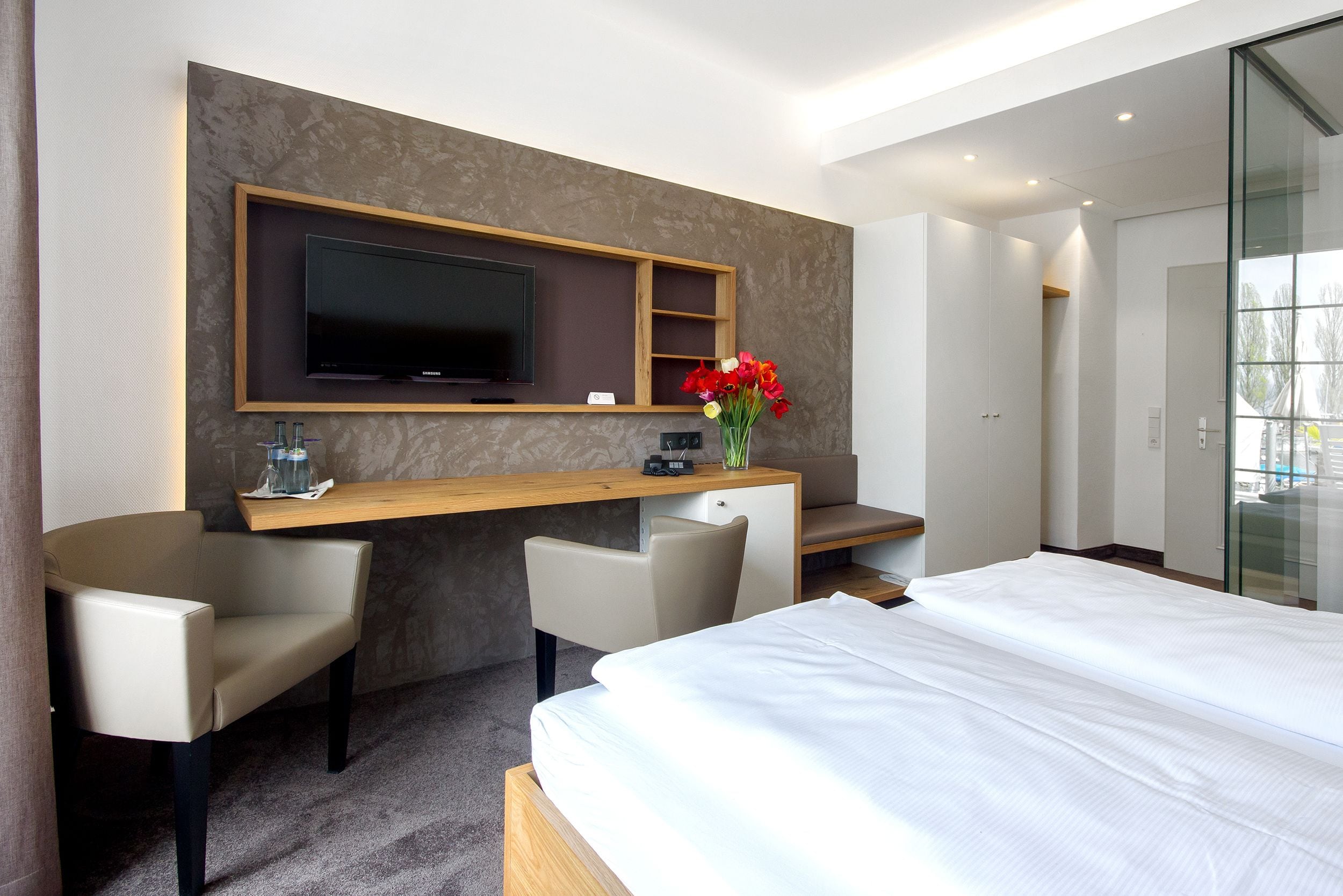 A modern hotel room with a large bed, a desk with a flat-screen TV, two chairs, and a vase with red flowers.
