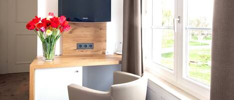 Ein Zimmer mit Schreibtisch aus Holz, einem Flachbildfernseher, einer Vase mit roten Blumen, einem Sessel und einem Fenster mit Blick ins Grüne.