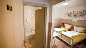 A hotel room with a wooden door marked '15', a small bathroom with a toilet and towel hanging, and a bed with a wooden frame and a yellow bedspread. - Hotel Gasthof Zur Linde (Hessdorf)