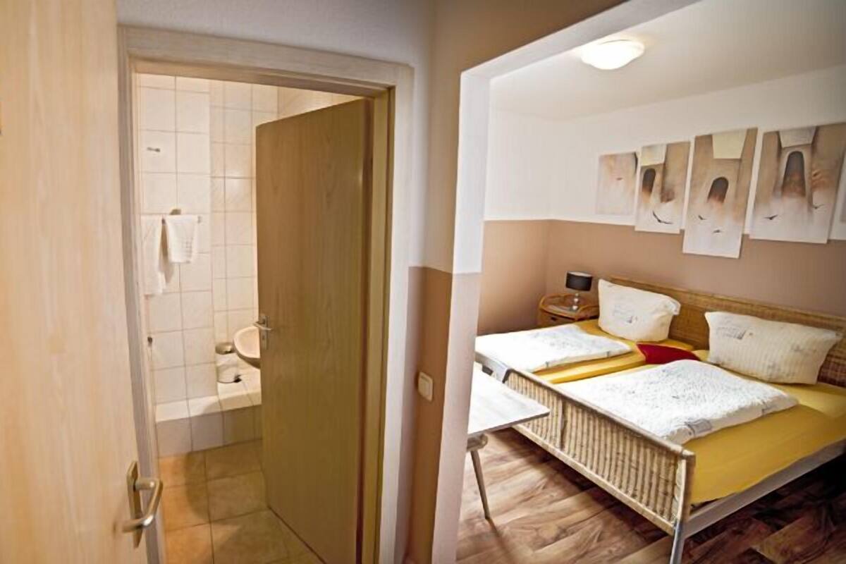 Una habitación de hotel con una puerta de madera marcada con el número 15, un baño pequeño con inodoro y toalla colgada, y una cama con estructura de madera y edredón amarillo.