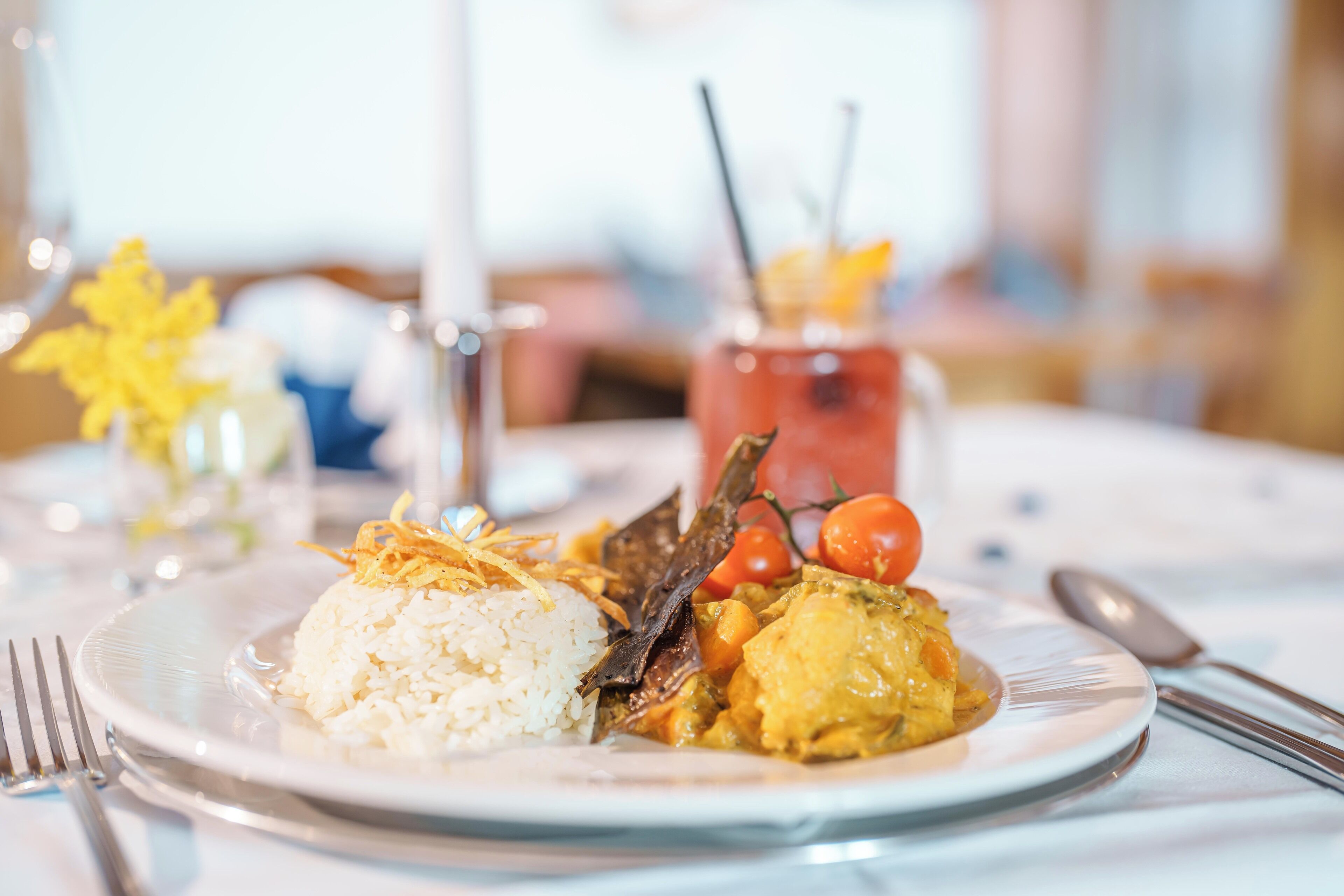 Une assiette de nourriture composée de riz et de curry, un verre contenant une boisson avec une paille, et une fourchette et une cuillère posées sur la table.