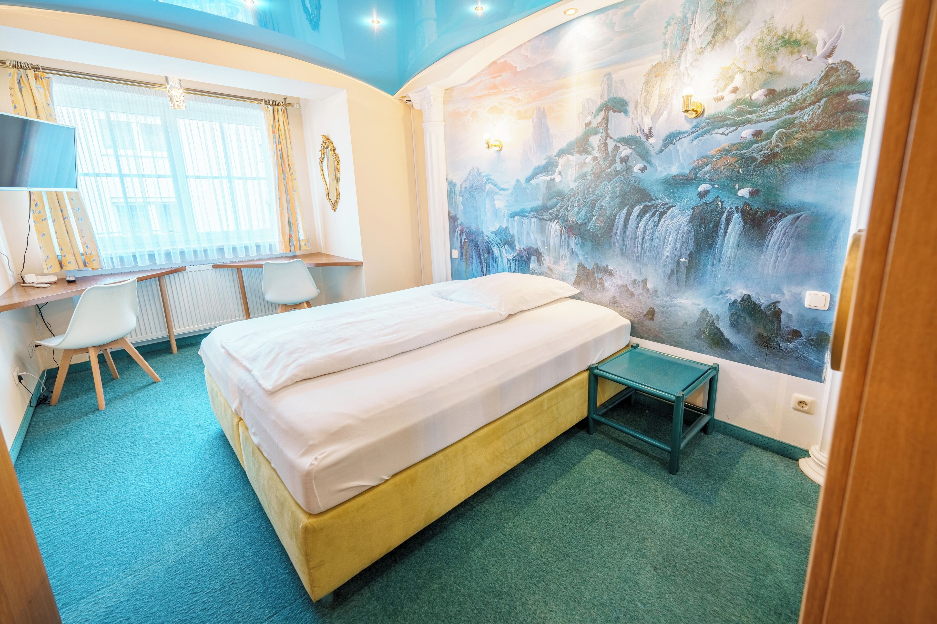 Habitación de hotel con cama, escritorio con silla, televisor y un mural de una cascada.