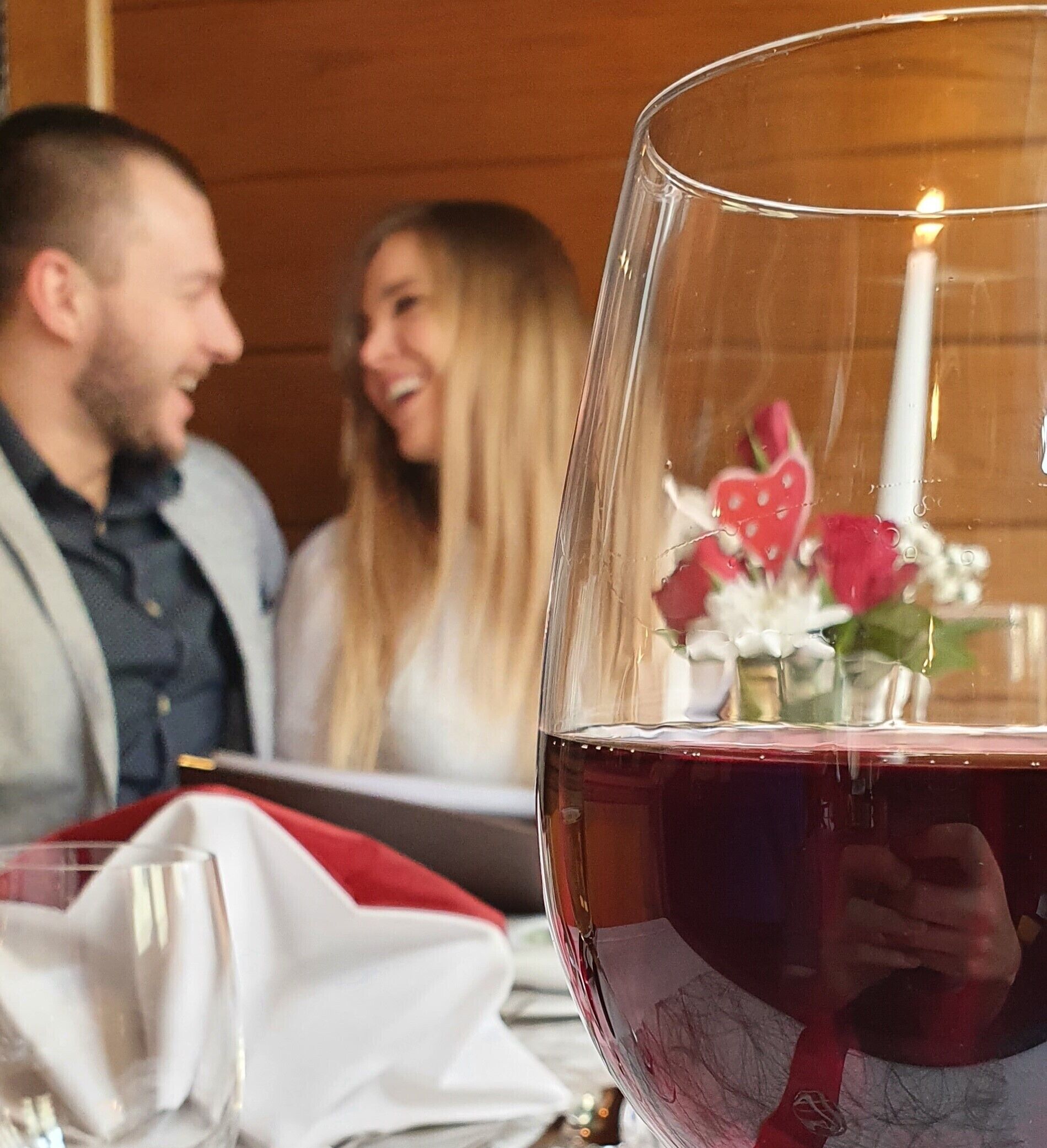 Primer plano de una copa de vino, una vela, un paño rojo y un jarrón pequeño con flores sobre una mesa.