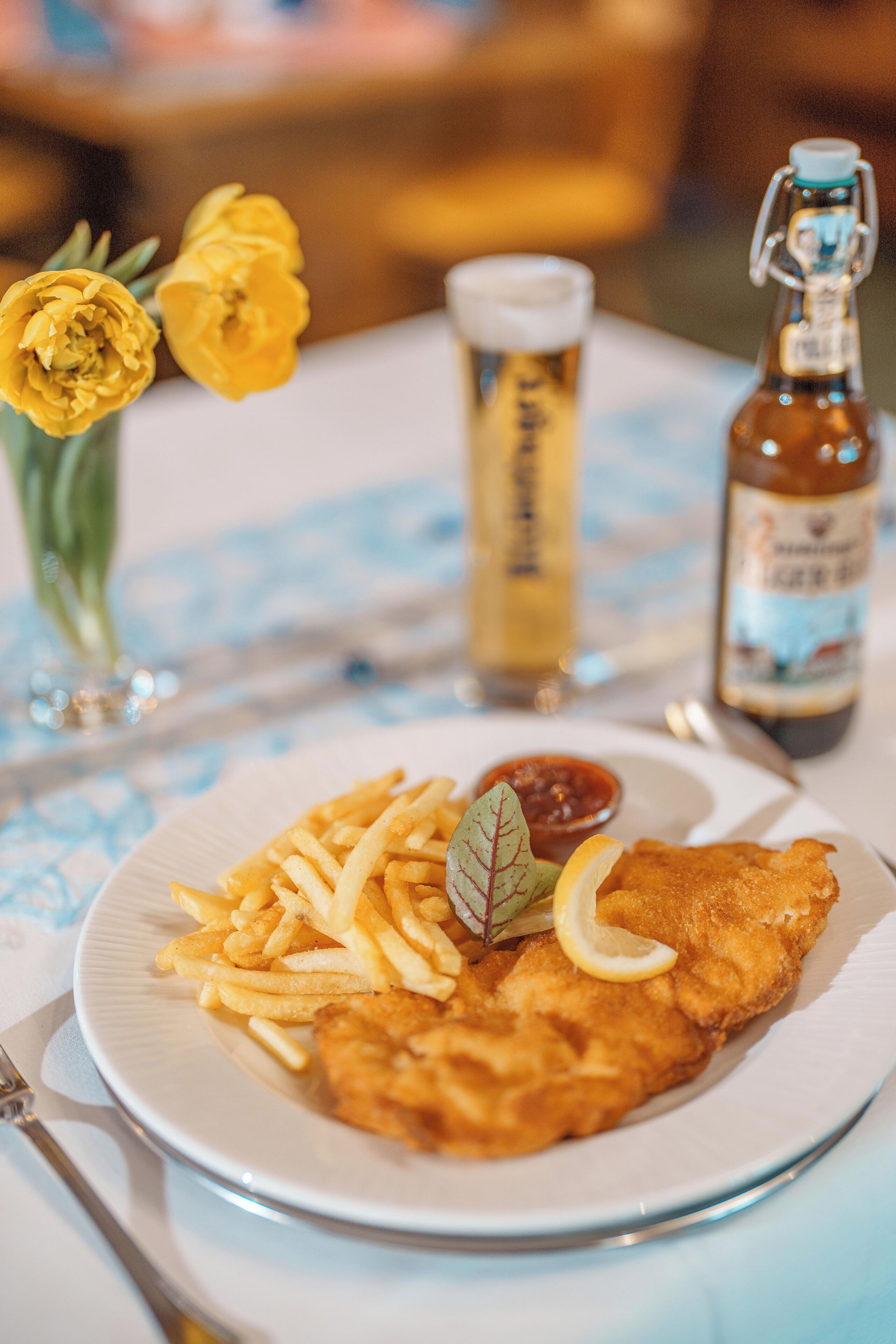 A plate of food with fries and a fish fillet, a glass of beer, and a bottle of beer on a table with a blue tablecloth.