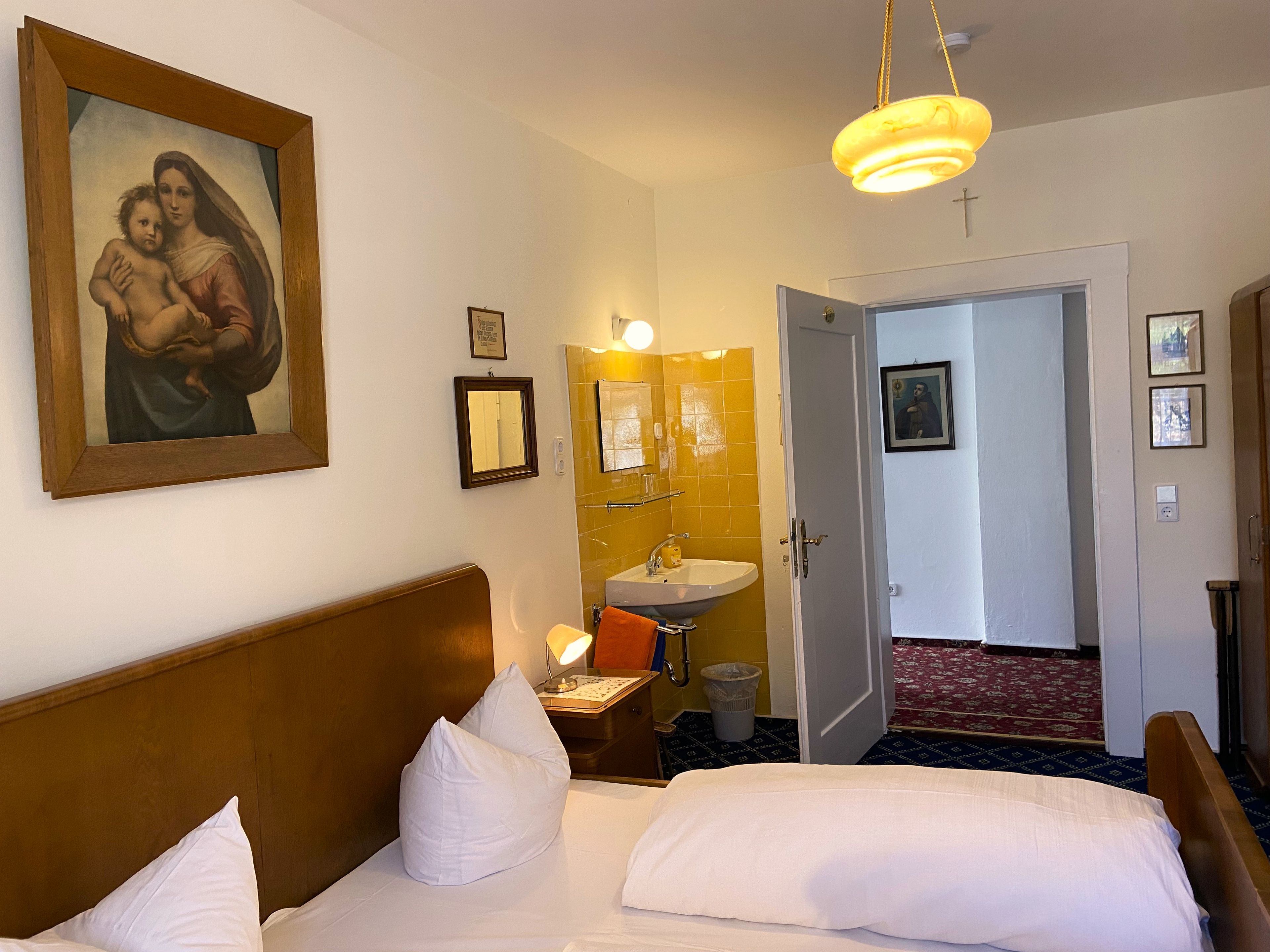 Una habitación de hotel con cama, un cuadro, un baño con lavamanos y espejo, y una puerta que da a otra habitación.