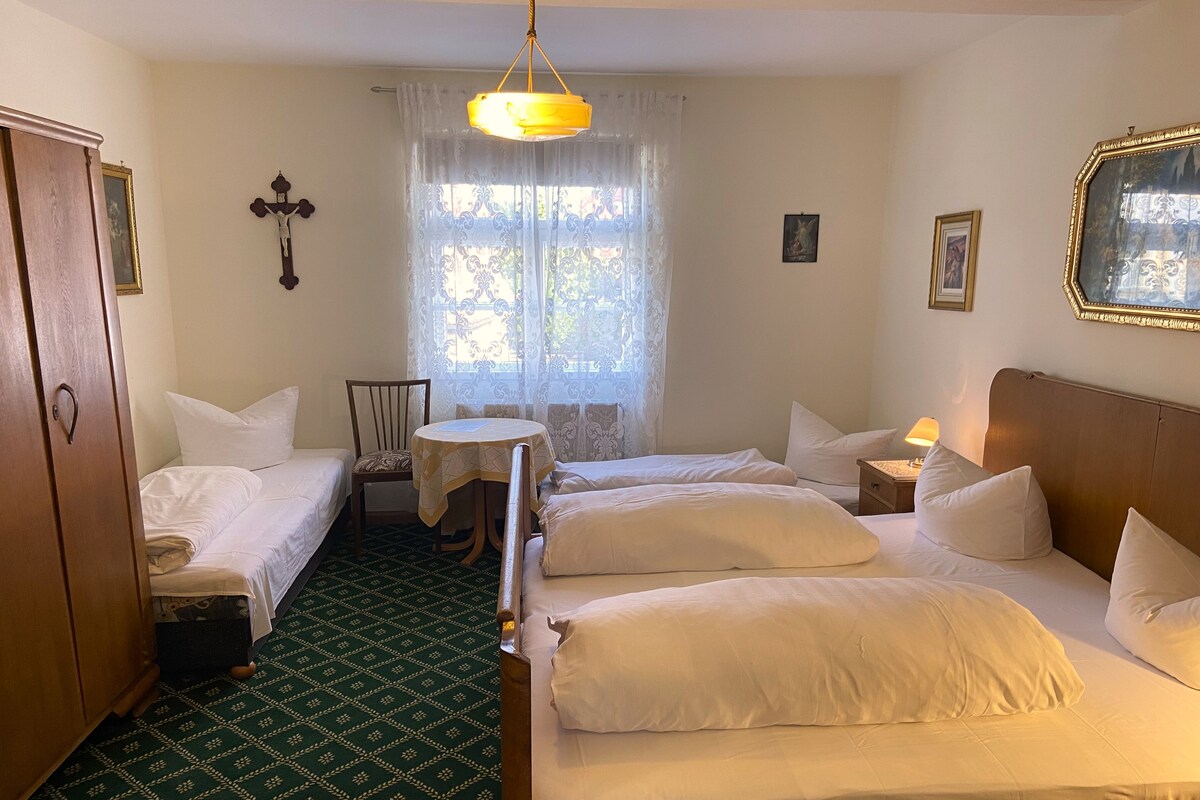 Habitación de hotel con dos camas, una mesita pequeña, una silla, un armario de madera, una cruz en la pared y una ventana con cortinas.