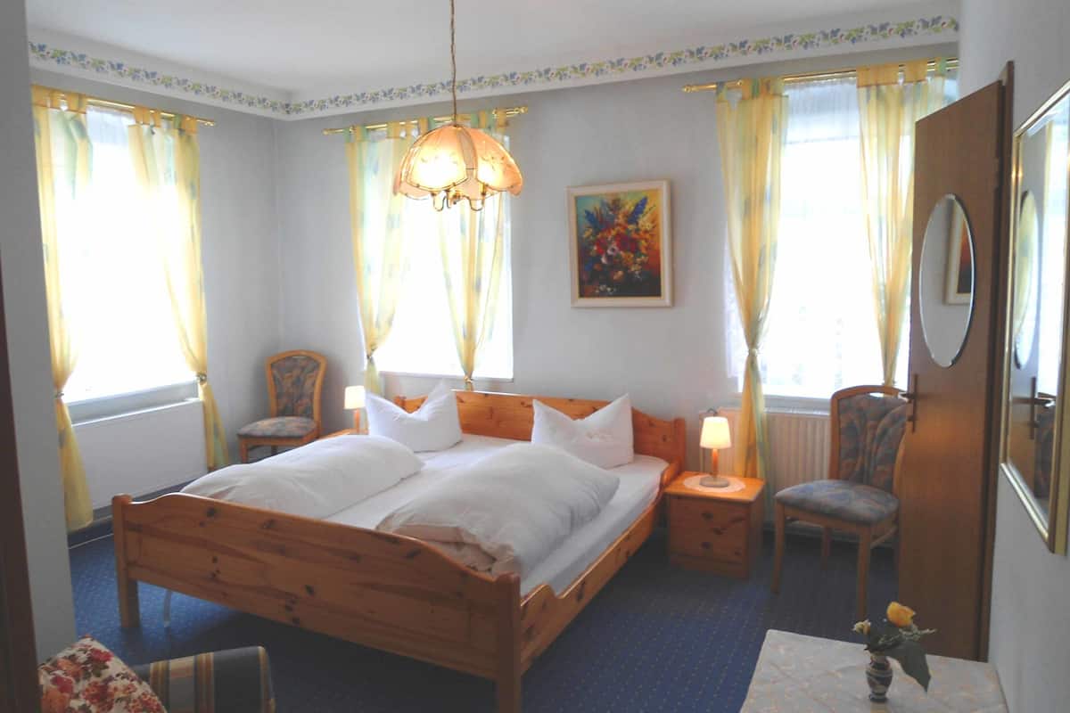 Un dormitorio con una cama de madera, dos mesitas de noche, una silla, una pequeña mesa con un florero y un cuadro en la pared.