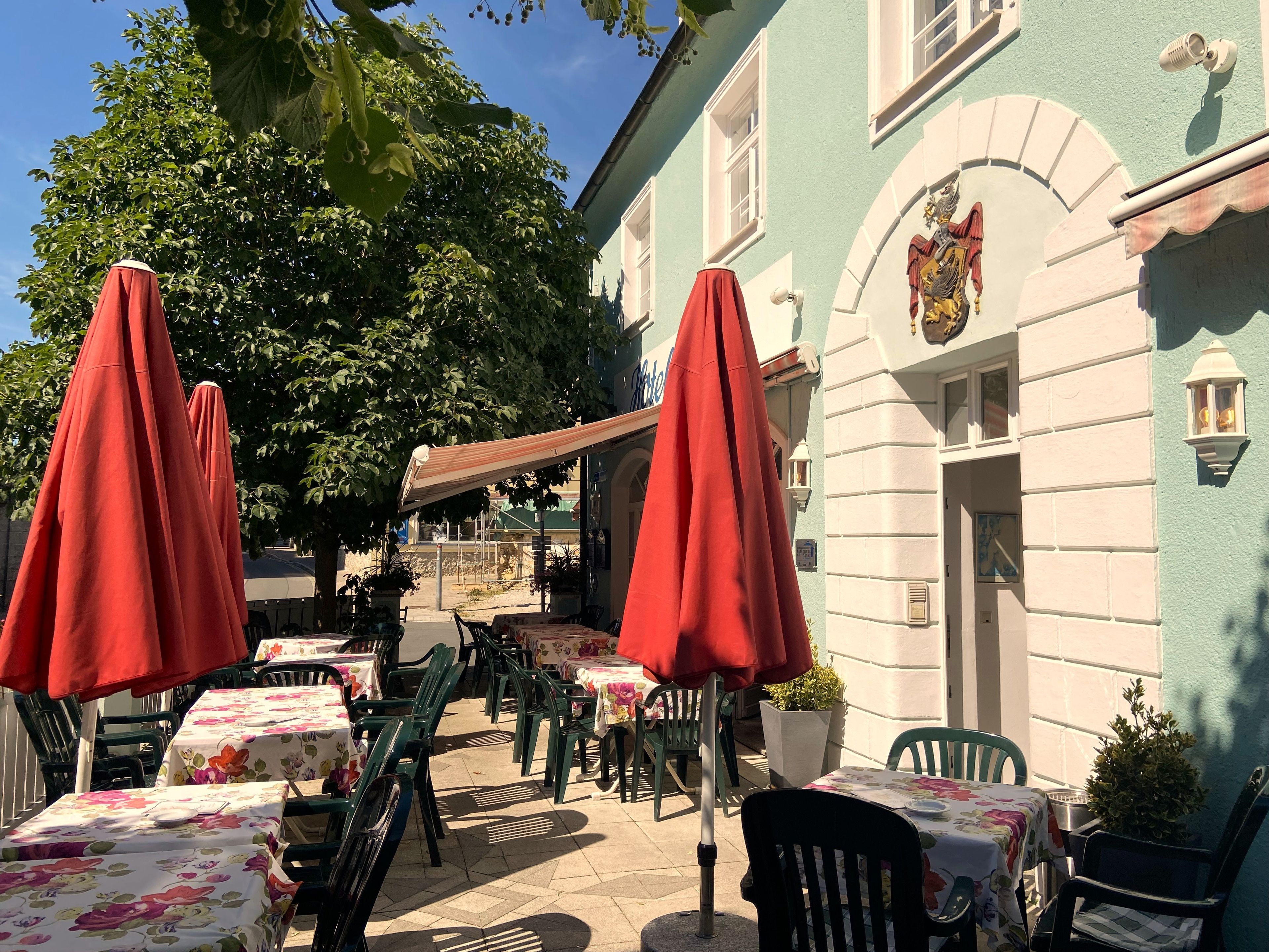 Un espace extérieur aménagé avec des tables et des chaises, des parasols rouges et un bâtiment orné d’un blason.