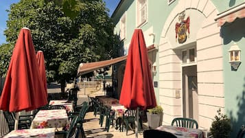 Un espace extérieur aménagé avec des tables et des chaises, des parasols rouges et un bâtiment orné d’un blason.