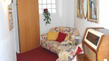 Una habitación pequeña y acogedora con un sofá floral, un aparador de madera con una radio y una ventana con vistas.