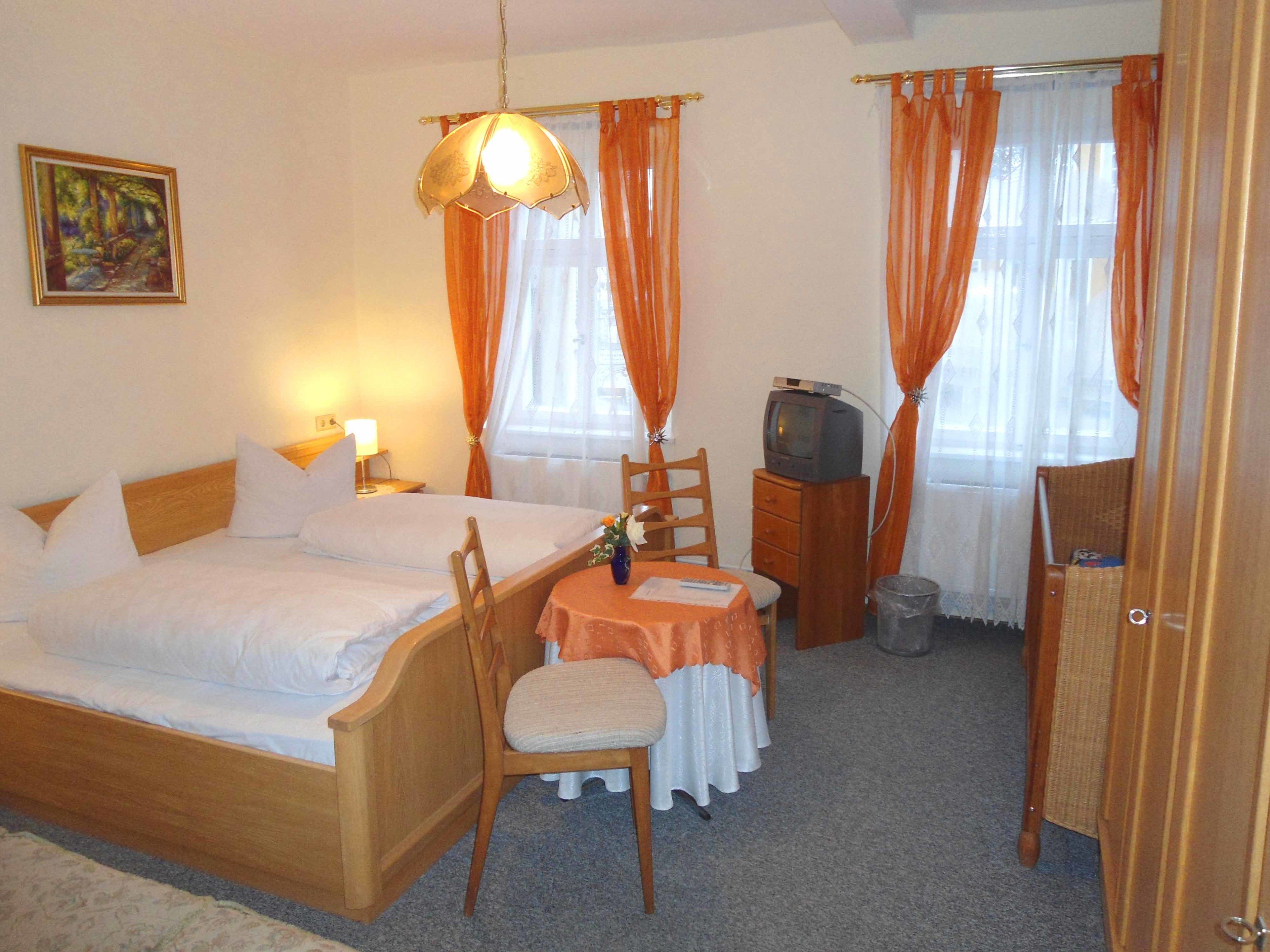 A hotel room with a wooden bed, a small table with a chair, a television on a wooden cabinet, and orange curtains.