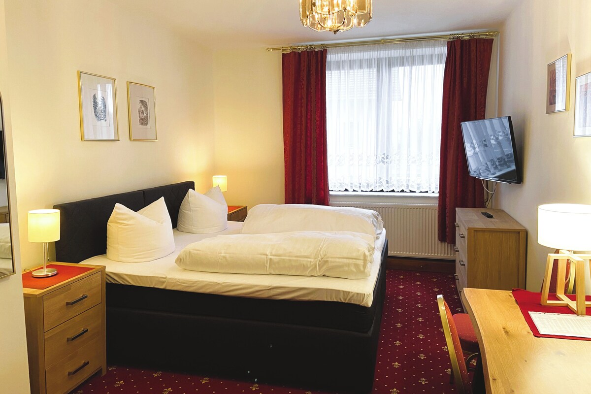 Una habitación de hotel con cama, mesitas de noche, escritorio, silla, televisor y ventana con cortinas.