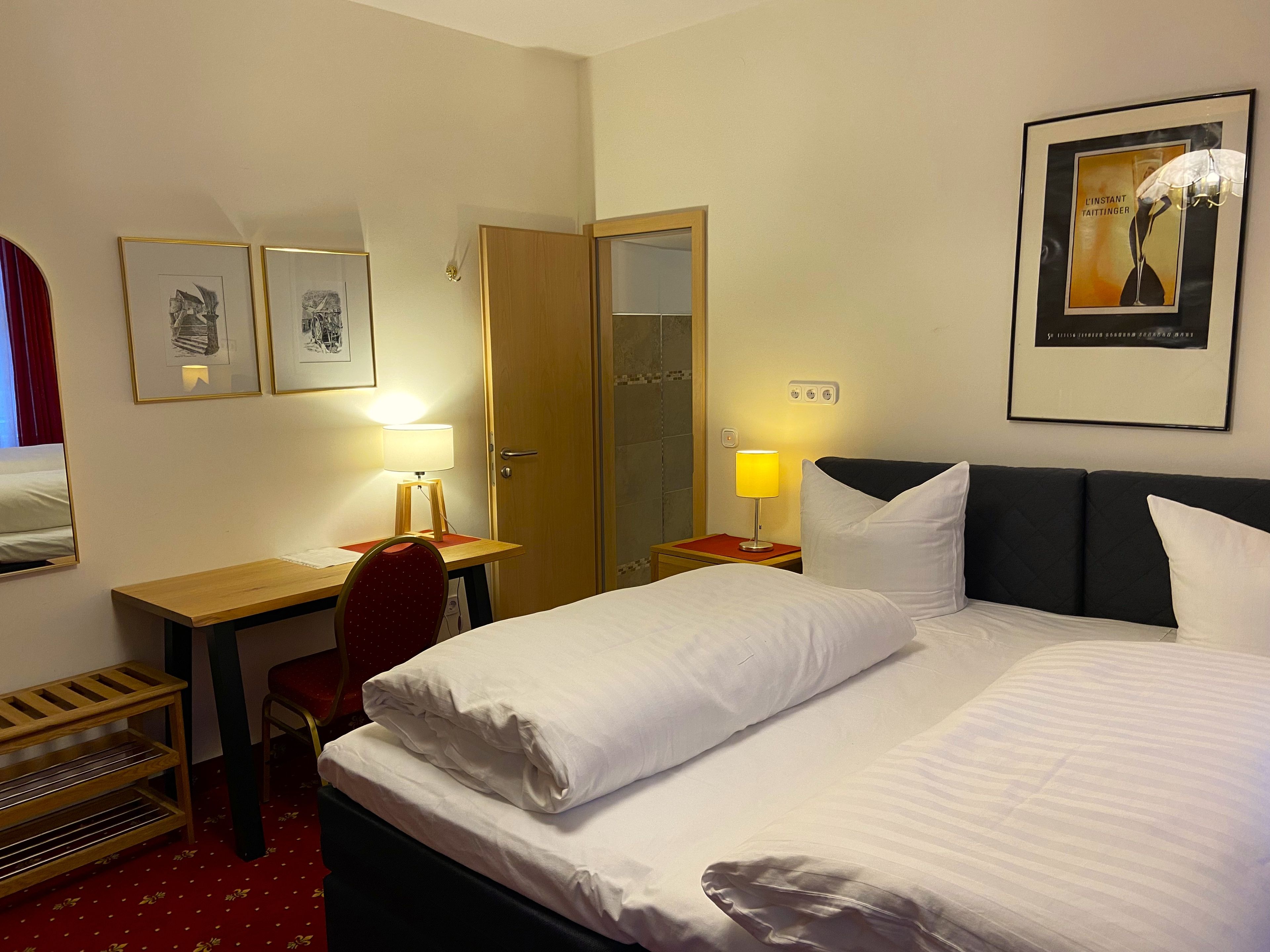 A hotel room with a bed, a desk, a chair, a bedside table, and a wall-mounted lamp.