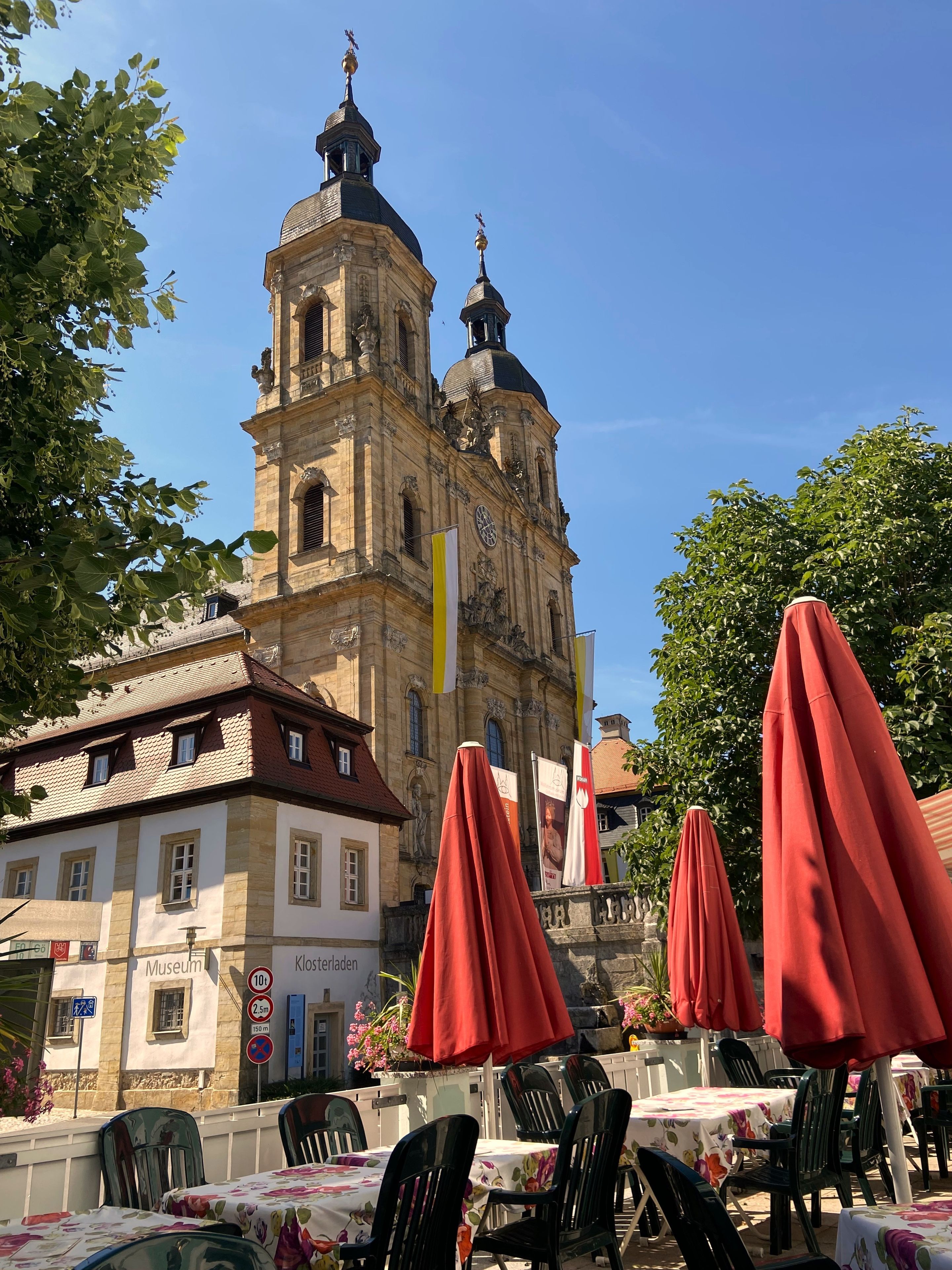 Ein historisches Gebäude mit einem Glockenturm, Außenplätzen mit roten Sonnenschirmen und einem Schild für ein Museum.