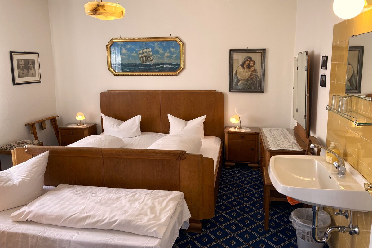Habitación de hotel con una cama grande, mesitas de noche con lámparas, un cuadro de un barco en la pared, un espejo y un baño con lavamanos e inodoro.